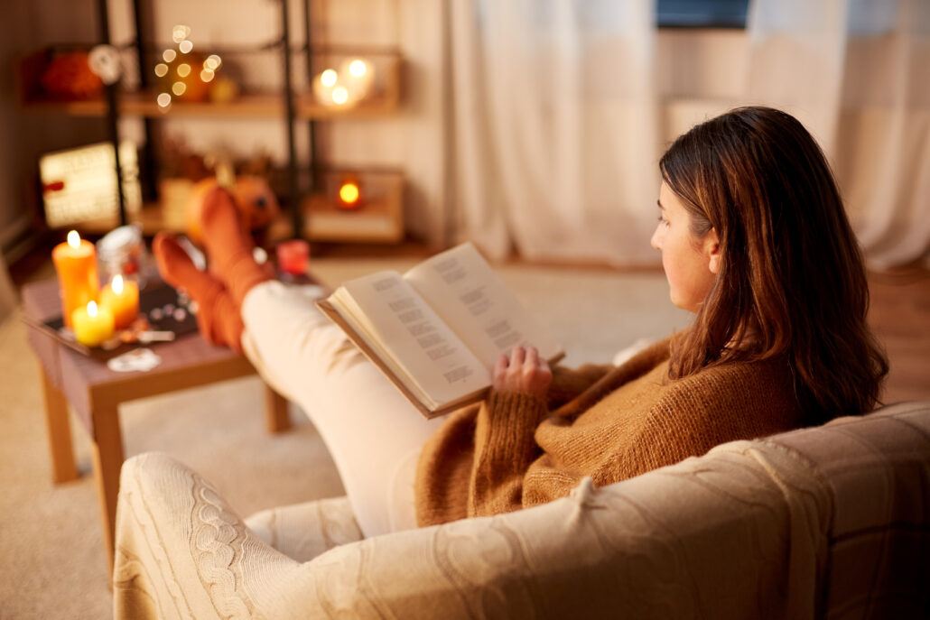Cozy woman reading a book on a sofa in a warmly lit living room with candles and autumn decor, creating a comfortable atmosphere for relaxing at home.